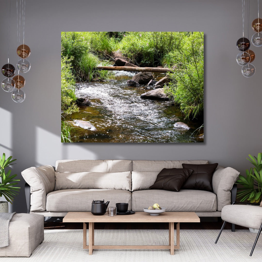 A extra large wall art print depicting a lush green stream with rocks and a fallen log in Wyoming, displayed above a modern beige sofa in a styled living room.
