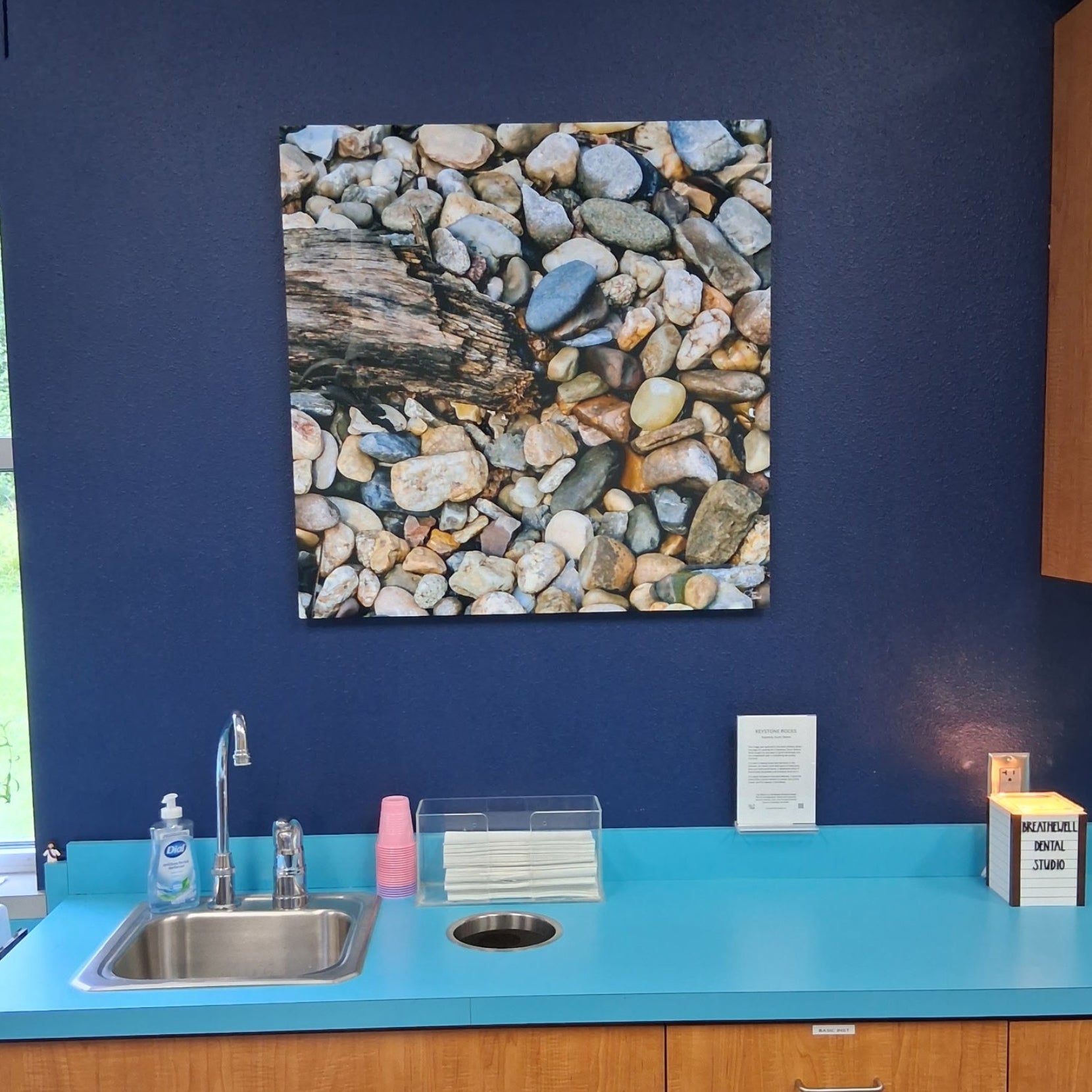 A square metal fine art print depicting a smooth multicolored pebbles and driftwood, from Keystone South Dakota - displayed on a dark blue dental office wall above a turquoise kitchenette counter with wood cabinets.