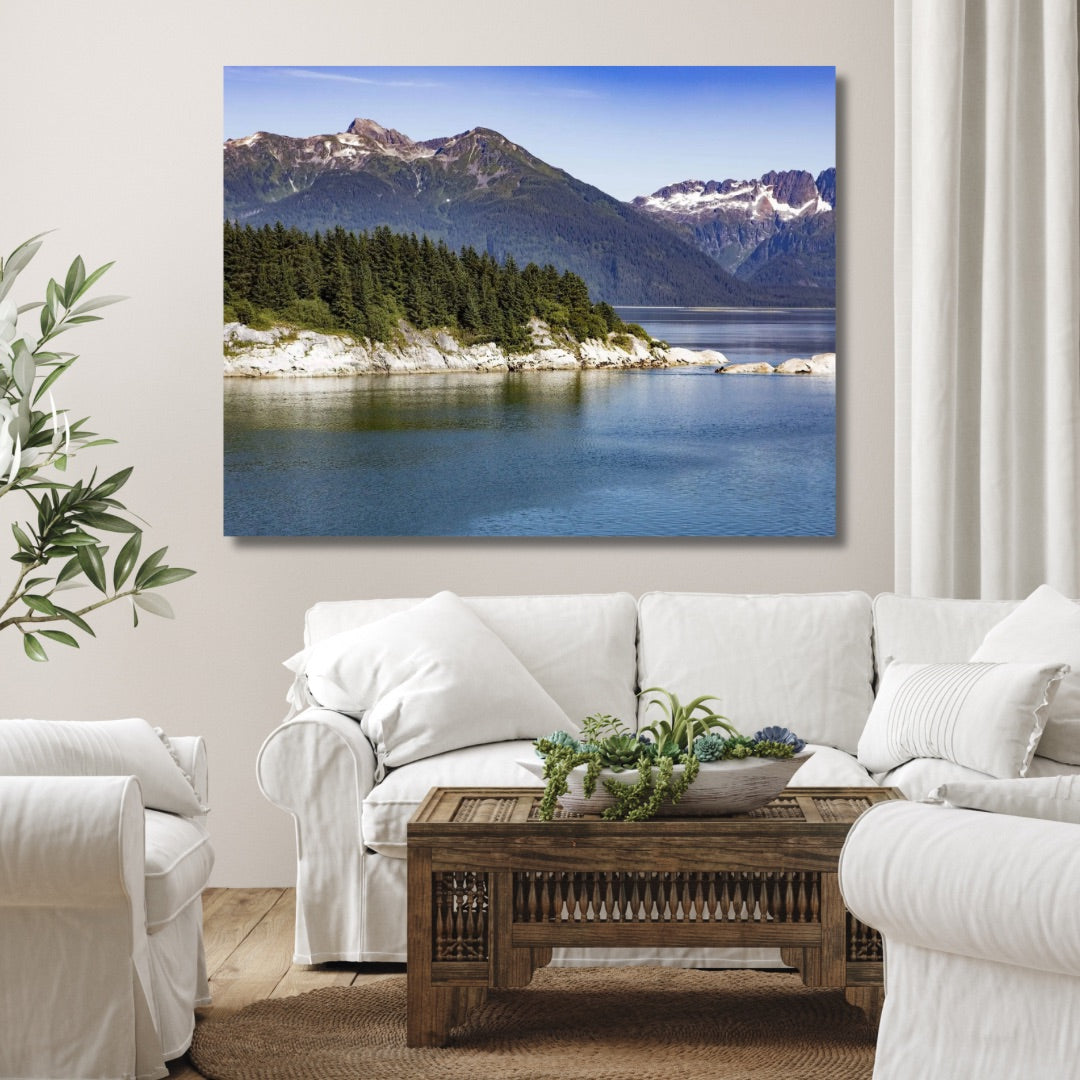 A framed wall art print showing a forest path of Glacier Bay Alaska with green trees and a wooden fence with sealions sunbathing on the rocks, displayed above a console table in a styled living room with a green armchair, books, and a woven basket.