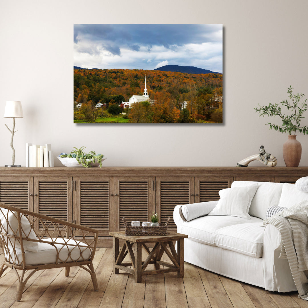 Stowe Vermont’s white church in the fall with orange leaves. Fine art photography on canvas is hanging above a light brown credenza in a casual setting. Could be traditional or farmhouse decor.