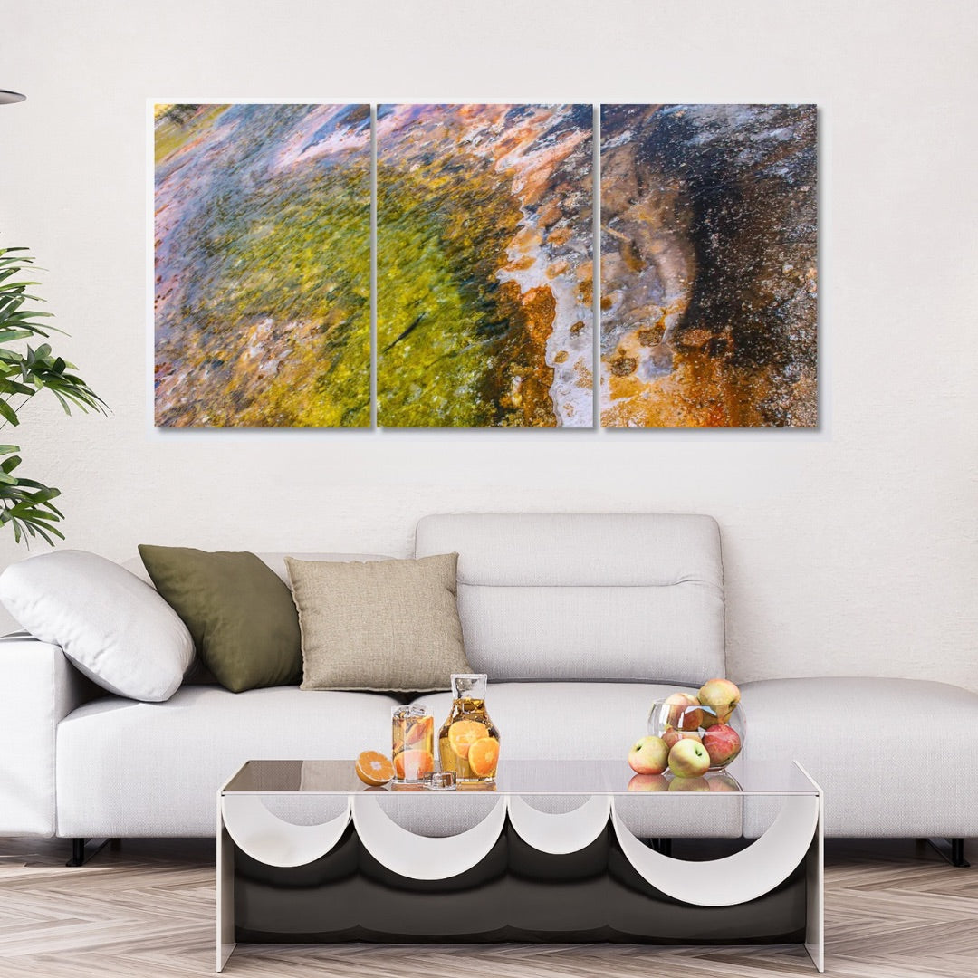 A three-panel abstract canvas wall art set hanging above a light gray sofa in a modern living room. The artwork features a textured, painterly design with green, gold, brown, and blue tones of the Devils Pool at Yellowstone National Park in Wyoming.