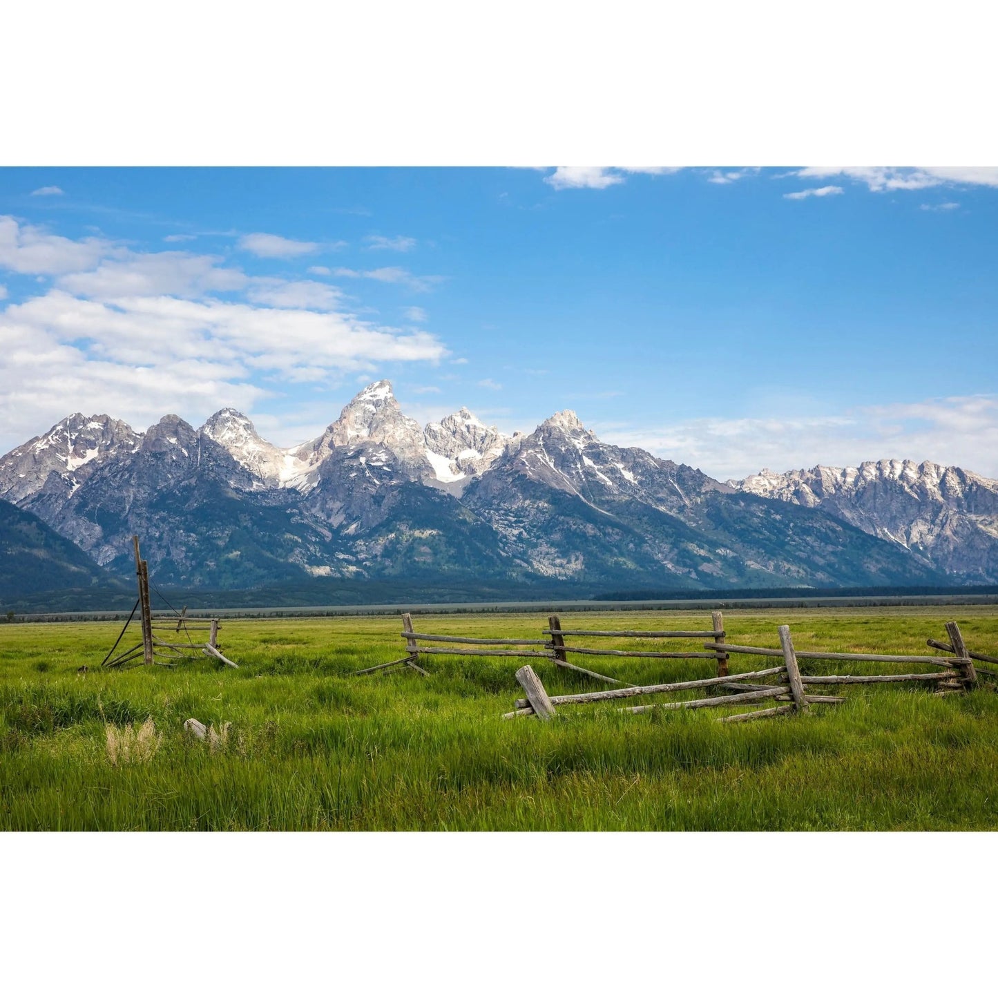 View of Grand Tetons from Mormon Row with green pasture and a wooden farmhouse fence