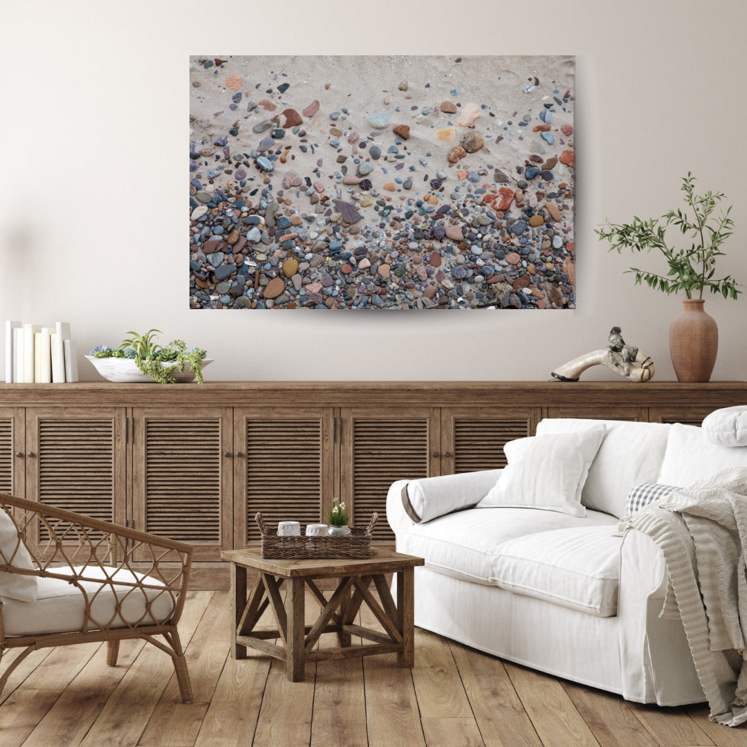 A cozy living room with wooden furniture, featuring a white couch and wicker chair. On the wall hangs a photo of colorful pebbles on sand. The space feels calm and inviting.