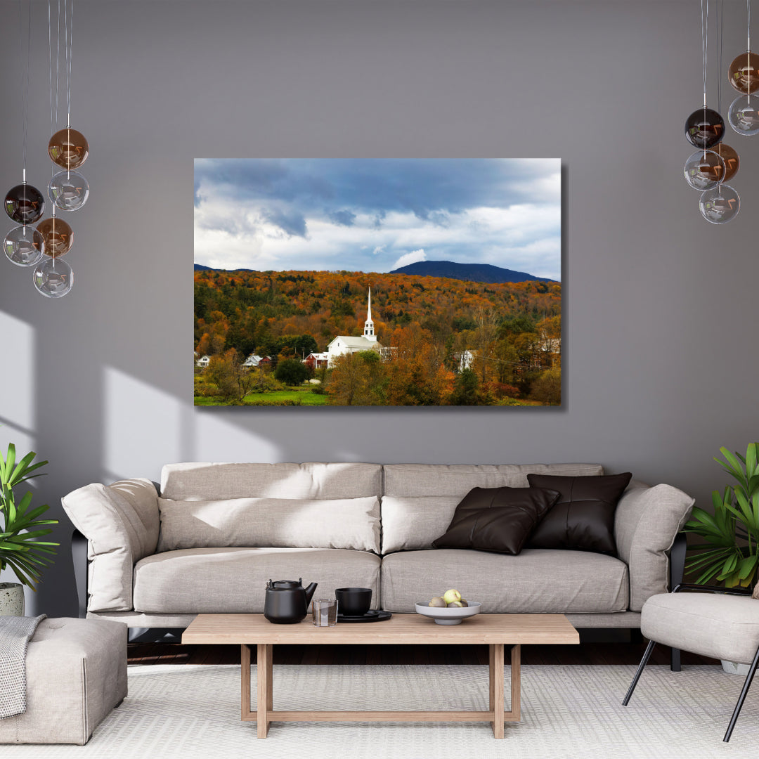 Stowe Vermont’s white church in the fall with orange leaves.  Large art hanging above a neutral colored couch with brown accented lights and pillows. 