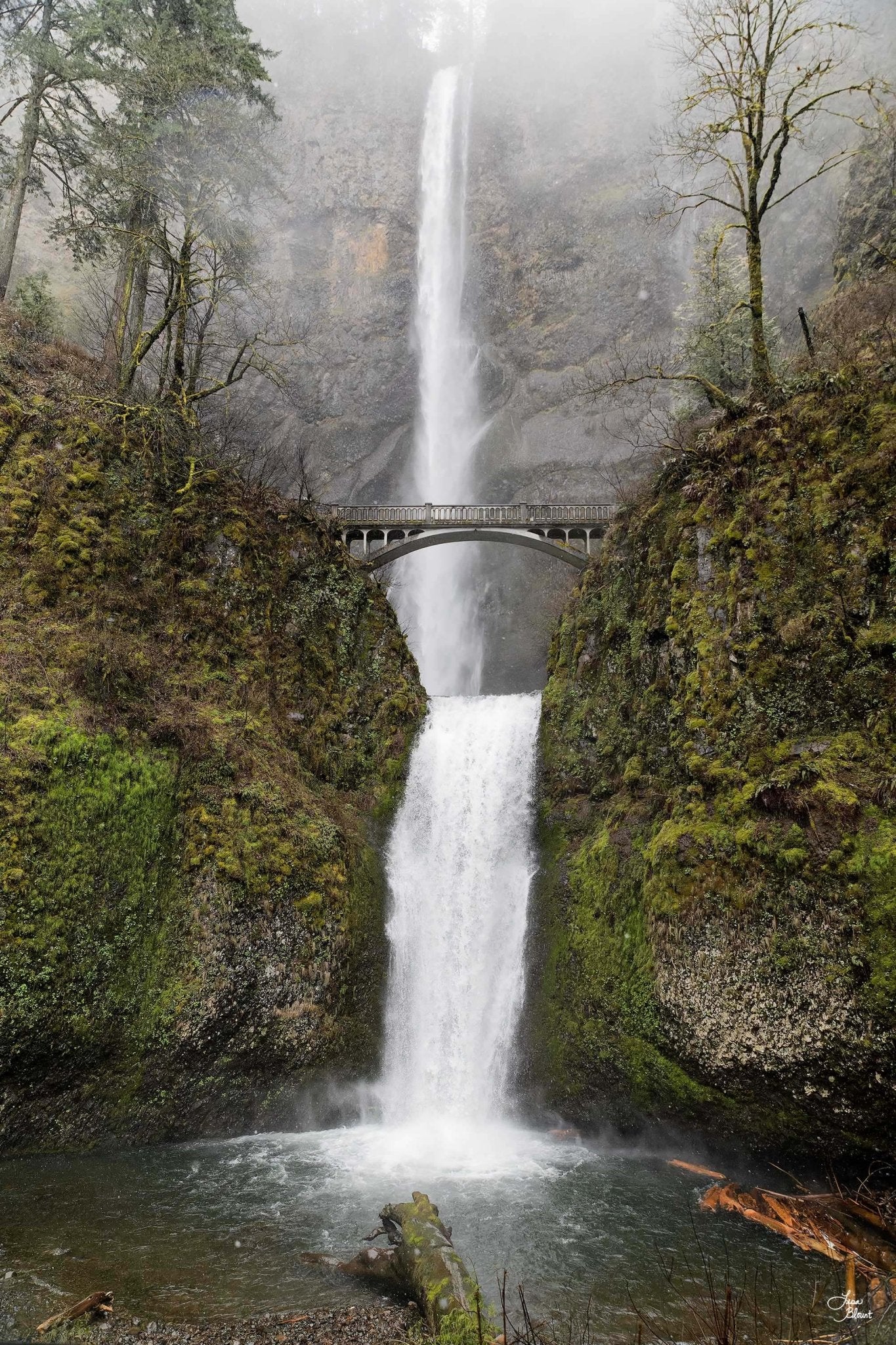 Fine art photograph of Multnomah Falls in Oregon, featuring the iconic stone footbridge, lush green moss, and mist rising from the double waterfall.